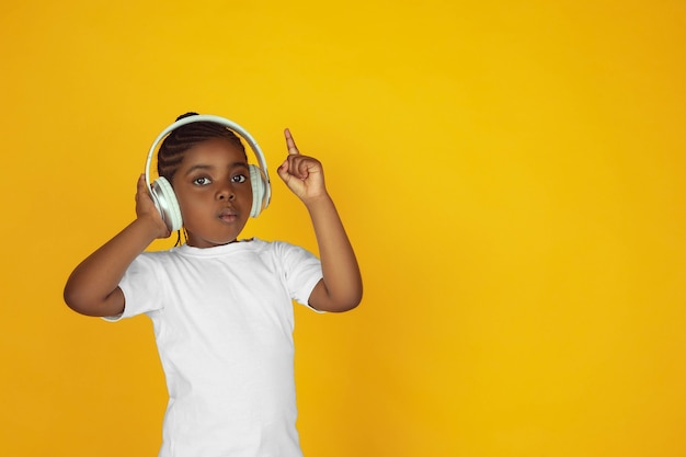 Photo pointant vers le haut, écouter de la musique. portrait de petite fille afro-américaine sur fond de studio jaune. enfant joyeux. concept d'émotions humaines, d'expression faciale, de ventes, d'annonces. espace de copie. ça a l'air mignon.