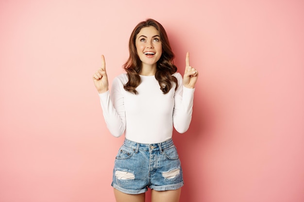 Pointant vers le haut. Heureuse fille élégante souriante, montrant l'annonce sur le dessus, démontrant la publicité, portant une tenue d'été élégante, fond rose.