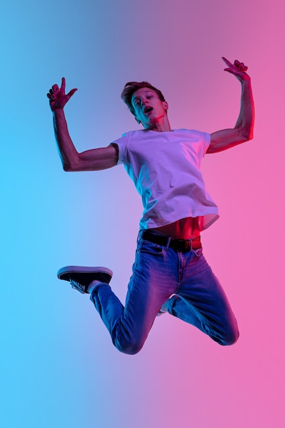 Pointant vers le haut. Jeune homme de race blanche sautant haut sur fond de studio dégradé bleu-rose à la lumière du néon. Concept de jeunesse, émotions humaines, expression faciale, ventes, publicité. Beau modèle en casual.