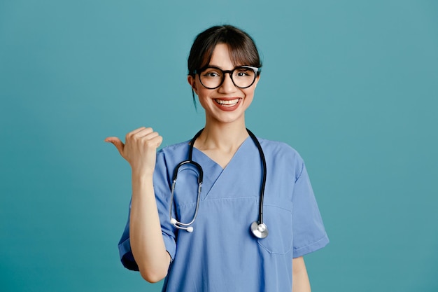 Points souriants à côté jeune femme médecin portant un stéthoscope fith uniforme isolé sur fond bleu