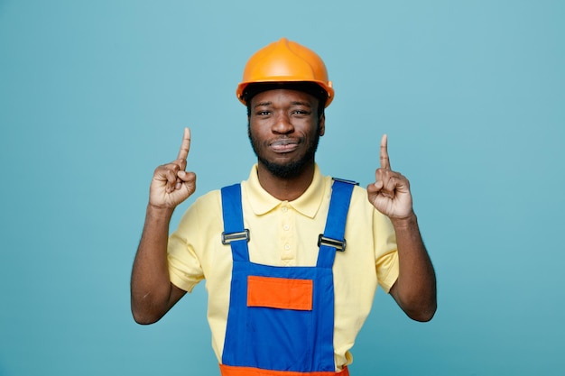 Points souriants vers le jeune constructeur afro-américain en uniforme isolé sur fond bleu