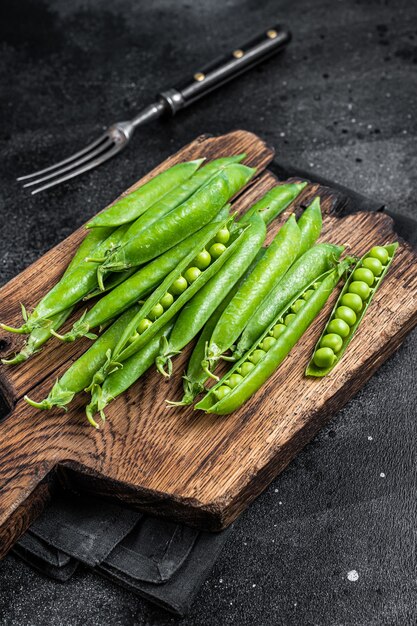 Pois frais et cosses de pois verts sur une planche à découper en bois. Fond noir. Vue de dessus.