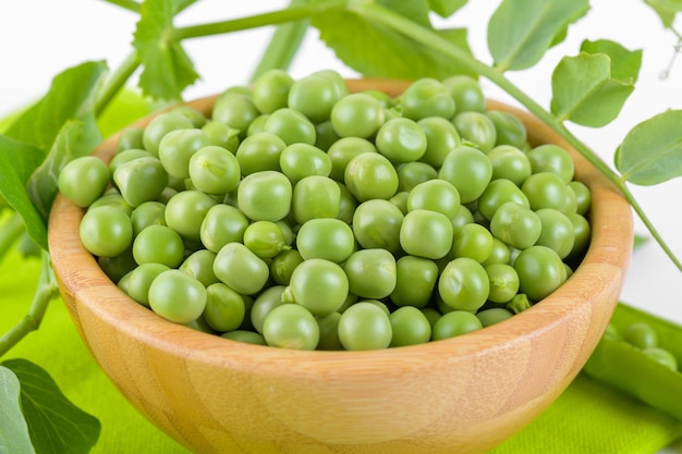 Pois verts frais dans un bol en bois avec des feuilles de plantes de pois sur une serviette verte sur fond blanc