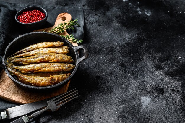 Poisson de capelan pané frit entier servi sur une poêle en métal. Fond noir. Vue de dessus. Espace copie