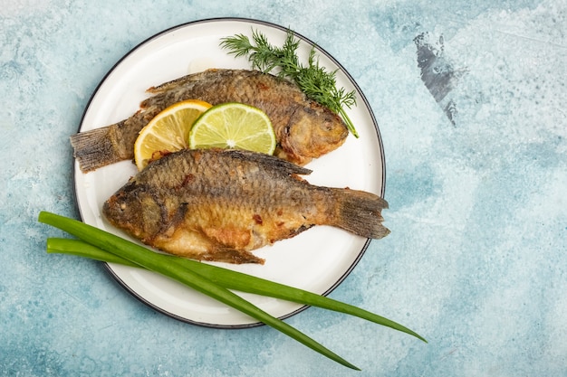 Poisson carpe frit sur une assiette avec des tranches de citron et de lime, avec des verts sur fond bleu Repas prêt. Copier l'espace