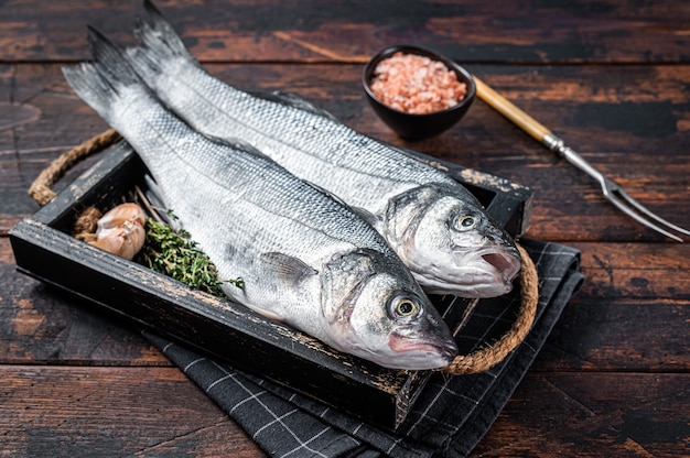 Poisson cru de bar ou bar dans un plateau en bois avec des herbes. Fond en bois sombre. Vue de dessus.