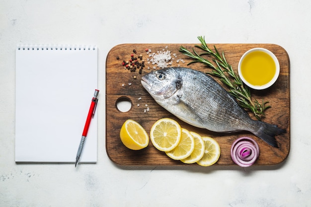 Poisson Dorado sur une planche à découper en bois rustique avec des épices et un cahier pour la recette ou le menu. Vue de dessus sur fond blanc.