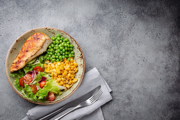 Poitrine de poulet au four sur assiette avec salade fraîche, pois verts et maïs, fond de pierre grise, vue de dessus. Repas fitness sain avec filet de poulet, équilibré en protéines et glucides. Frais généraux, copiez l'espace