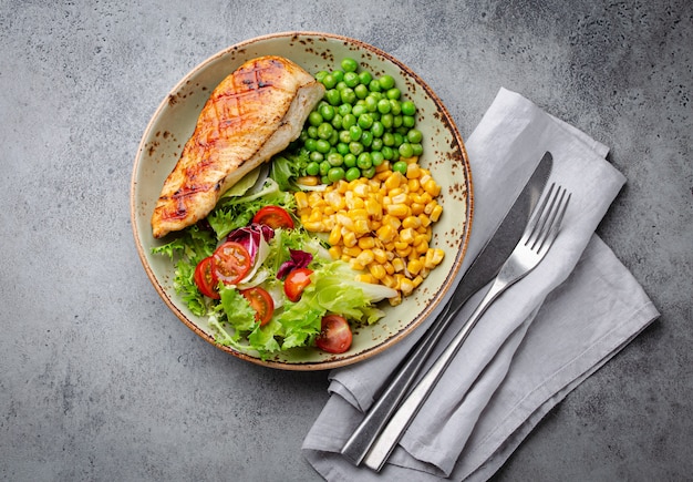 Poitrine de poulet au four sur assiette avec salade fraîche, pois verts et maïs, fond de pierre grise, vue de dessus. Repas fitness sain avec filet de poulet, équilibré en protéines et glucides. Régime et nutrition