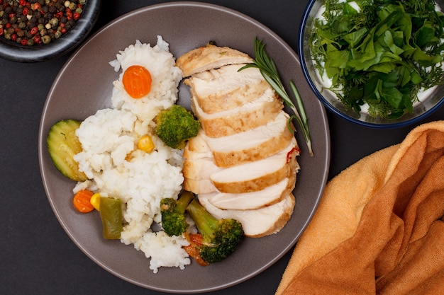 Poitrines de poulet au four ou filet avec riz, légumes et légumes verts sur assiette, bols en verre avec sauce et baies de piment de la Jamaïque sur fond noir. Vue de dessus.