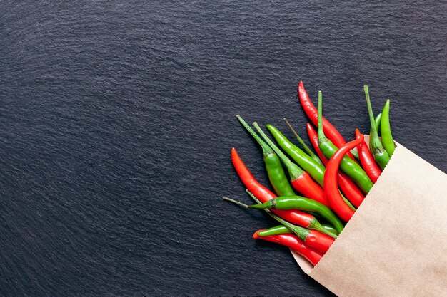 Poivron rouge et vert épicé frais dans un sac en papier kraft sur une ardoise, divers piments colorés et poivrons de Cayenne sur fond sombre d'en haut. Vue de dessus, copiez l'espace.