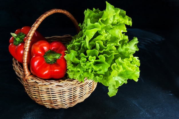 Poivrons rouges mûrs et bouquet de salade verte dans un panier en osier sur fond sombre.