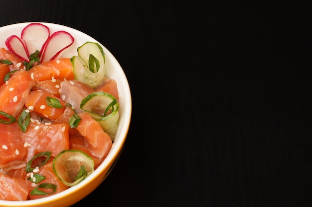 Poke hawaïen au saumon et graines de sésame, concombre frais, radis et oignons verts dans un bol en porcelaine sur fond noir. Vue de dessus avec espace de copie. Fruits de mer biologiques