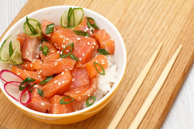 Poke Hawaïen Au Saumon Et Graines De Sésame, Riz Bouilli, Concombre Frais, Radis Et Oignons Verts Dans Un Bol Avec Des Baguettes En Bois Sur Une Planche à Découper En Bois