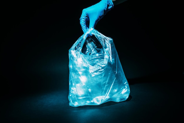 Pollution plastique. Pollution des déchets. Problème d'écologie. Main volontaire dans un gant bleu prenant des bouteilles usagées collectées dans un sac en cellophane isolé sur fond sombre.