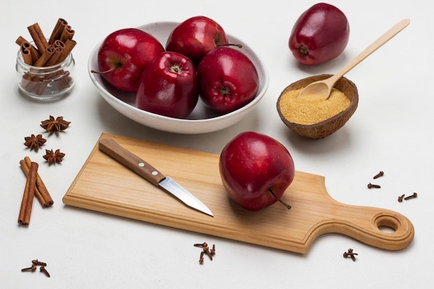 Pomme et couteau sur une planche à découper Pommes dans un bol Bâtons de cannelle dans un bocal en verre Cassonade dans une coque de noix de coco Vue de dessus Fond blanc