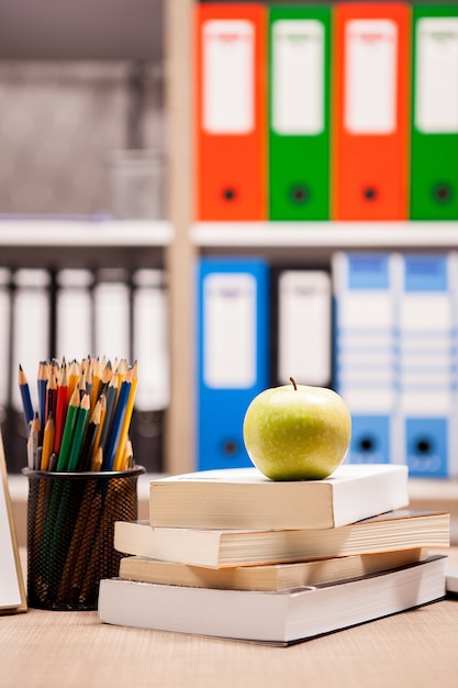 Pomme verte sur une pile de livres à côté d'un cahier et de crayons sur une table avec un tableau blanc flou à l'arrière. Notion d'école