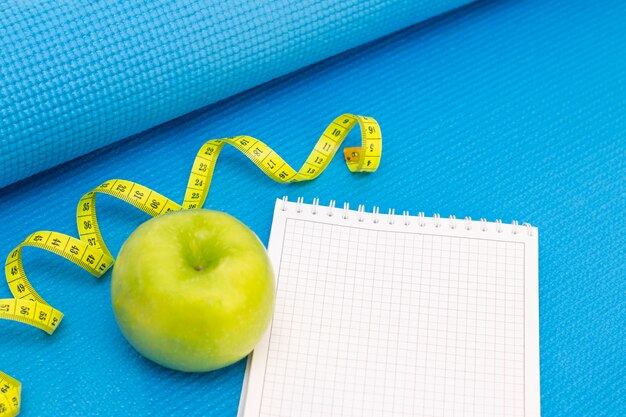 Pomme verte, ruban à mesurer, cahier sur un tapis de sport de fond de couleur bleue. préparation pour la saison estivale et la plage, perte de poids et concept sportif