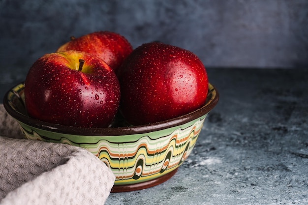 Pommes rouges fraîches juteuses avec des gouttes d'eau dans un bol peint sur une serviette de cuisine sur fond gris