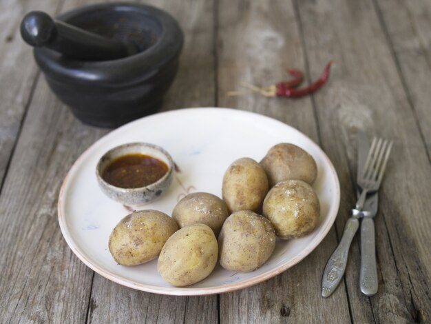 Photo pommes de terre bouillies dans leur peau. entier avec poivron rouge, sel et sauce épicée sur assiette blanche, vieille table en bois, style rustique. gros plan, copiez l'espace.