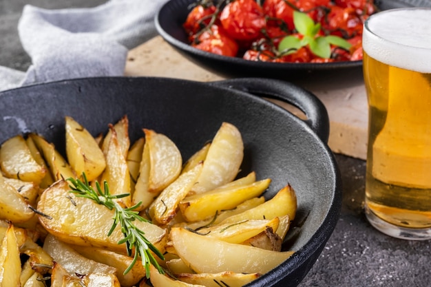 Pommes de terre rôties au romarin en cocotte en fer et assiette de tomates confites sur fond sombre.