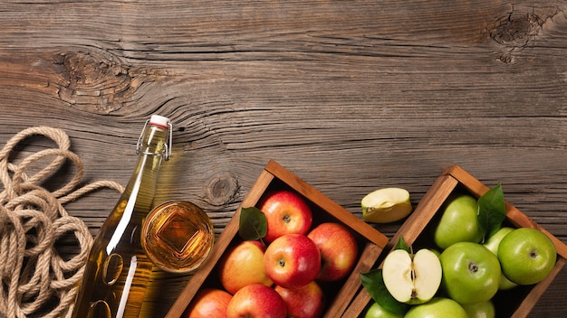 Pommes vertes et rouges mûres dans une boîte en bois avec branche de fleurs blanches, verre et bouteille de cidre sur une table en bois. Vue de dessus avec un espace pour votre texte.