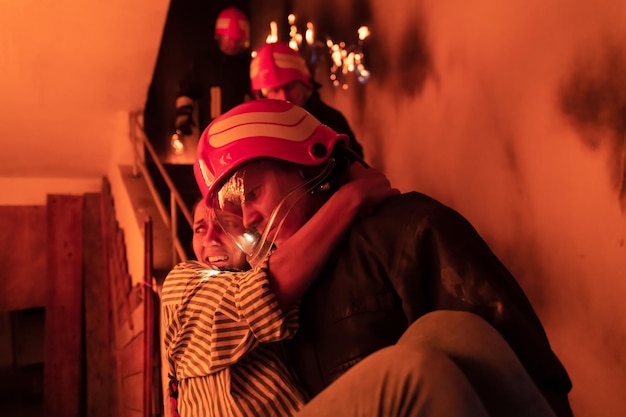 Un pompier courageux descend les escaliers d'un immeuble en feu et tient une fille sauvée dans ses bras. Ouvrez le feu et un pompier en arrière-plan. Photo de haute qualité