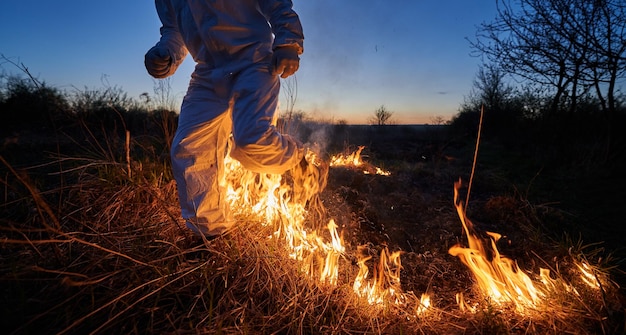 Pompier écologiste éteignant le feu dans le champ la nuit Vue recadrée de l'homme en combinaison de protection contre les radiations qui traverse l'herbe brûlante avec de la fumée Concept de catastrophe naturelle