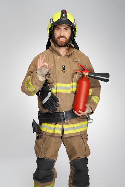 Pompier heureux avec extincteur montrant un signe correct à l'intérieur, vue de face du pompier barbu dedans
