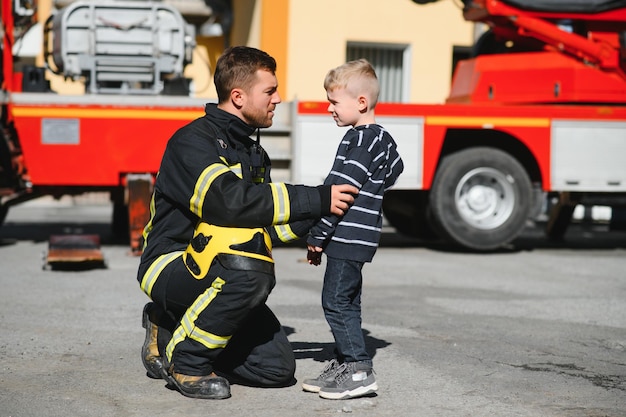 Un pompier prend un petit enfant garçon pour le sauver Voiture de pompier sur fond Pompier avec enfant dans ses bras Concept de protection