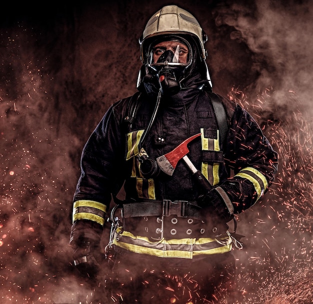 Un pompier vêtu d'un uniforme et d'un masque à oxygène tient une hache rouge debout dans des étincelles de feu et de la fumée sur un fond sombre.