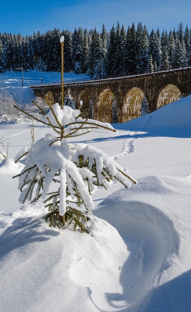 Pont en arc de viaduc en pierre sur le chemin de fer à travers la forêt de sapins enneigés de montagne Congères de neige sur le bord de la route et de givre sur les arbres et les fils électriques