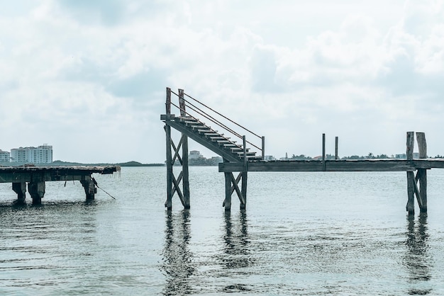 Pont jetée en bois abandonné et endommagé avec escalier au-dessus de la surface de l'eau de mer contre ciel nuageux