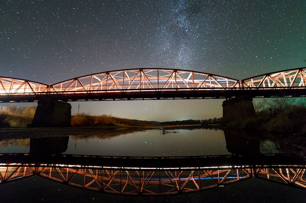 Pont métallique éclairé sur des supports en béton réfléchi dans l'eau sur un ciel étoilé sombre avec la constellation de la voie lactée. Concept de photographie de nuit.