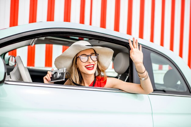 Poortrait d'une jeune femme heureuse sortant de la fenêtre de la voiture avec appareil photo sur le fond du mur rouge pendant les vacances d'été