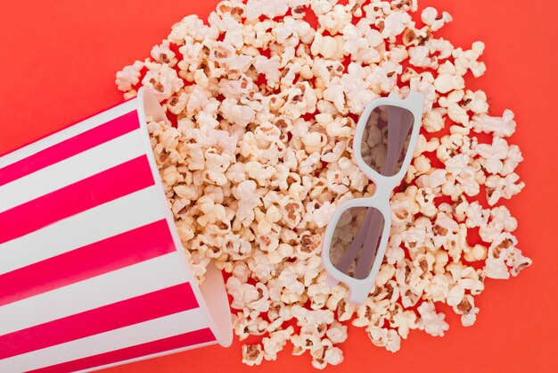 Pop-corn versant d'un bol en papier sur un fond rouge, et des lunettes 3d pour regarder le film, une vue de dessus