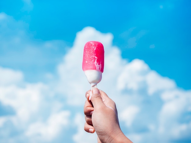 Popsicle rose et blanc dans la main de la femme. Main féminine tenant un popsicle rose fondant sur fond de ciel bleu et de nuages en été.