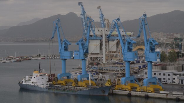 Port industriel avec grues à conteneurs et cargo amarré, ville côtière éloignée et montagnes en arrière-plan