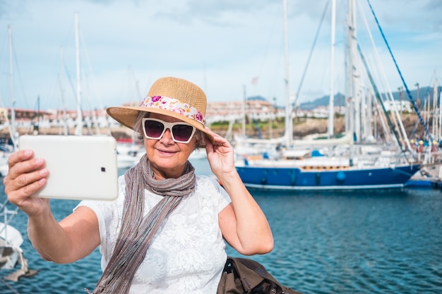 Un port avec yachts et voiliers. Une femme âgée aux cheveux gris prend un selfie avec le paysage avec sa tablette. Journée nuageuse et venteuse