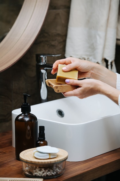 Porte-savon en bois dans les mains d'une femme mûre avec un bloc de savon écologique. Elle est debout devant le lavabo d'une salle de bain.