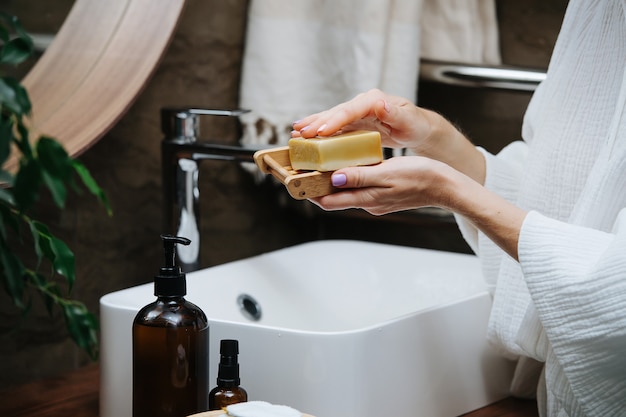 Porte-savon dans les mains d'une femme mûre avec un bloc de savon écologique. Elle est debout devant le lavabo d'une salle de bain.