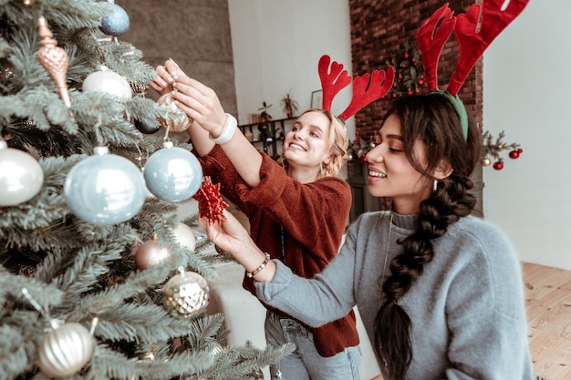 Porter des oreilles de cerf. Joyeuses jeunes filles mettant des jouets de Noël sur l'arbre vert dans le salon avant les vacances à venir