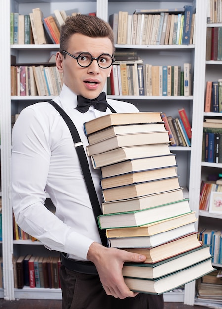 Porter une pile de livres. Jeune homme choqué en chemise et noeud papillon portant une lourde pile de livres et regardant la caméra