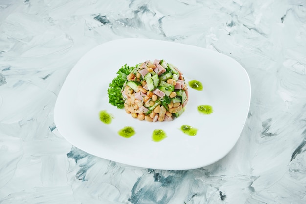 Portion de salade fraîche et saine avec des haricots, du jambon et des champignons marinés, assaisonnée de pesto sur une plaque blanche. Table grise.