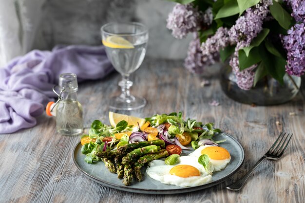 Une portion de salade de maïs avec des oignons rouges, des tomates jaunes, des asperges et deux œufs sur une assiette sur une table en bois.