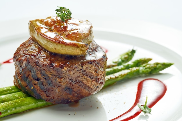 Portion de steak mignon grillé au foie gras et aux asperges, sauce aux baies dans une assiette blanche. Isolé sur une surface grise.