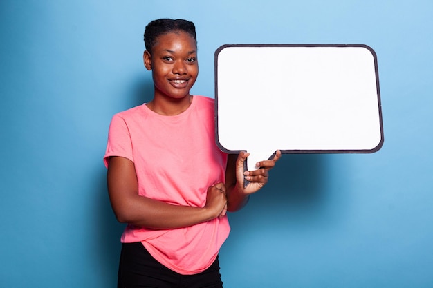 Portrait d'un adolescent afro-américain joyeux souriant à la caméra tout en tenant une bannière de texte vide debout en studio avec un fond bleu. Notion de publicité. Concept de tableau blanc