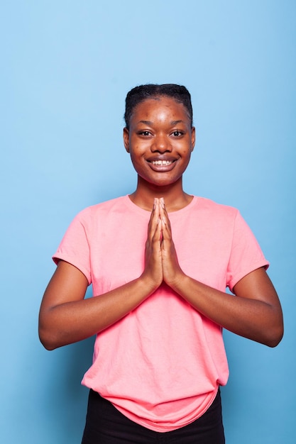 Portrait d'un adolescent afro-américain religieux priant Dieu pour demander de la gratitude debout en studio avec fond bleu. Jeune femme tenant les paumes ensemble dans la prière. Notion de religion