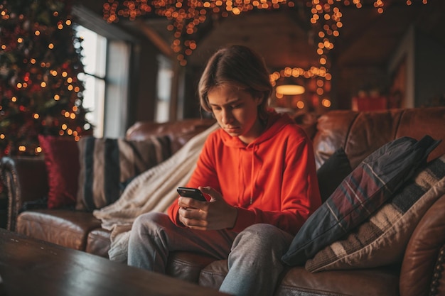 Portrait d'un adolescent beau garçon souriant et authentique à l'aide d'un téléphone portable à l'intérieur de la maison de Noël