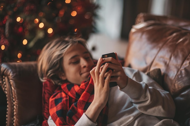 Portrait d'un adolescent beau garçon souriant et authentique à l'aide d'un téléphone portable à l'intérieur de la maison de Noël